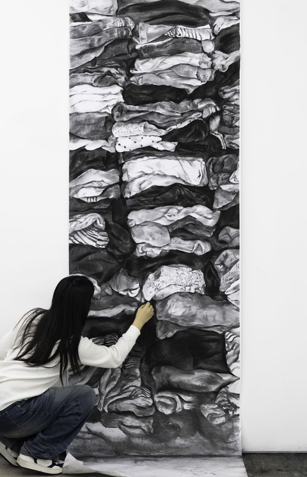 Esoo crouching beside a floor-to-ceiling charcoal drawing of stacked folded clothes