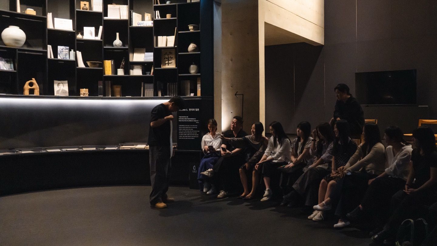A speaker addressing a seated group beside shelves of ceramics and books